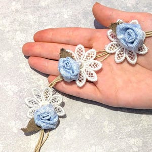 May include: A decorative headband featuring three light blue fabric roses, each centered on a white lace flower motif. The roses are attached to a thin, natural-colored twine cord, accented with small brown fabric leaves at the base of each flower.