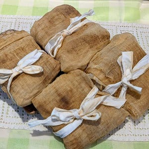 May include: Four rectangular bundles of tan, textured, semi-sheer fabric, each individually wrapped and secured with a white fabric tie knotted in the center. The bundles have a slightly distressed, woven appearance with visible fibers and frayed edges.
