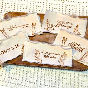 May include: Five rectangular off-white fabric strips with frayed edges, each featuring brown printed text and botanical illustrations. The text includes "With God All Things Are Possible", "Count Your Blessings", "JOHN 3:16", "Faith", and "Let your little light shine!".
