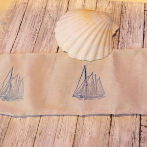 May include: A cream-colored fabric strip with blue sailboat illustrations. A large, white seashell rests on top. The fabric is placed on a weathered, light-colored wooden surface, creating a nautical theme.