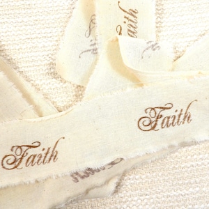 May include: White fabric ribbon with the word "Faith" written in brown cursive on it. The ribbon is folded and layered, creating a textured look.