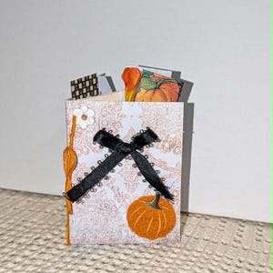 May include: A small rectangular paper card featuring a light orange damask pattern, a black ribbon tied in a cross shape, a small white flower embellishment, and an orange pumpkin illustration, with additional paper inserts visible at the top.