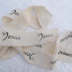 May include: White fabric ribbon with the word "Jesus" printed on it multiple times. The ribbon is frayed and has a rustic look.