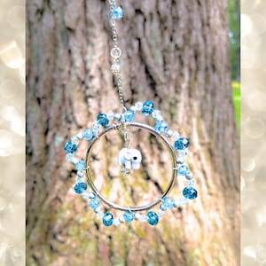 May include: A circular silver-toned metal hoop ornament featuring a dangling white elephant charm in the center. The hoop is decorated with a perimeter of alternating light blue, dark blue, and clear faceted beads on a silver-toned chain.