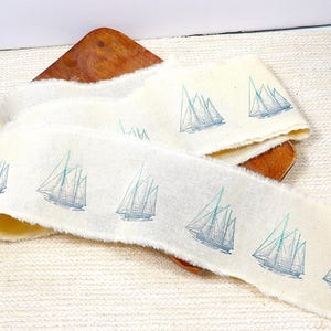May include: A long strip of off-white fabric with raw, frayed edges, featuring a repeating pattern of blue sailboats printed along its length. The fabric has a textured, woven appearance and is draped in a loose, folded arrangement.