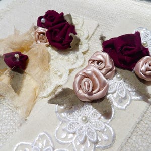 May include: Close-up of handmade floral embellishments. The designs feature burgundy velvet and blush satin roses, accented with pearls and lace. The fabric flowers are arranged on cream-colored fabric.