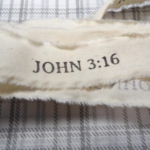 May include: Close-up of a cream-colored fabric ribbon with frayed edges. The text "JOHN 3:16" is printed in black ink across the ribbon. The background features a gray and white plaid pattern.