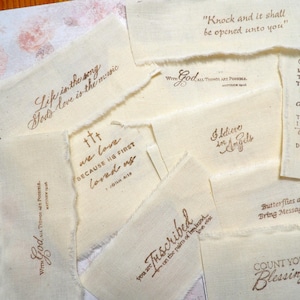 May include: A collection of off-white fabric strips with frayed edges, each featuring brown printed text with various inspirational and religious quotes including "Knock and it shall be opened unto you," "Life is the song," and "Count your blessings."