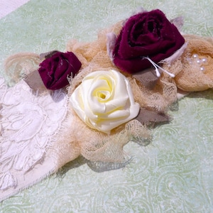 May include: A handmade floral accessory featuring fabric roses in burgundy and cream, accented with pearls and lace. The design includes a cream-colored lace base, beige netting, and decorative buttons. The arrangement is set against a light green patterned background.