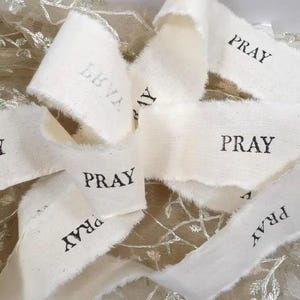 May include: Close-up of off-white fabric ribbons with frayed edges. The word "PRAY" is printed in black on several ribbons. Some ribbons have the word "LOVE" printed on them. The ribbons are arranged on a textured background.