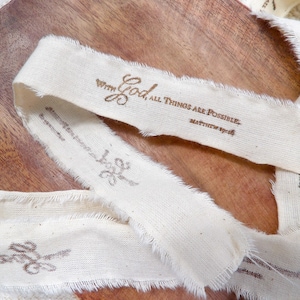 May include: Cream-colored fabric ribbon with frayed edges, printed with the words "With God, ALL THINGS ARE POSSIBLE" and "Matthew 19:26" in brown ink. The ribbon is lying on a wooden surface.