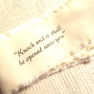 May include: A cream-colored fabric strip with frayed edges, featuring the quote "Knock and it shall be opened unto you" in dark brown script. The fabric rests on a textured, off-white surface.