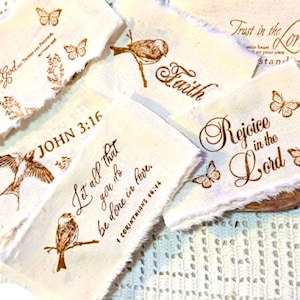 May include: A collection of rectangular off-white fabric scraps with frayed edges, each featuring brown ink prints of butterflies, birds, and religious text including "Faith," "John 3:16," "Rejoice in the Lord," and "Let all that you do be done in love."