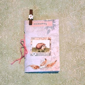 May include: A handmade journal with a light blue and floral patterned cover. The journal features a small illustration of a hedgehog and the word "memories". A pink ribbon and a brown clip with a white flower accent the journal.