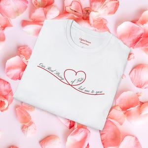 May include: White t-shirt with a crew neck featuring a red heart and the text "Our Red String of Fate led me to you" in a script font. The shirt is surrounded by pink rose petals.