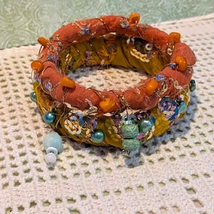 May include: A layered bangle bracelet featuring a top band wrapped in textured orange fabric with orange beads and a bottom band wrapped in mustard yellow fabric. The piece is embellished with metallic wire, sequins, and various blue and green beads.