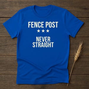 Peut inclure: Un t-shirt bleu roi avec le texte "FENCE POST" au-dessus de trois étoiles et les mots "NEVER STRAIGHT" en blanc. Le t-shirt est présenté sur une surface en bois avec une tige de blé.