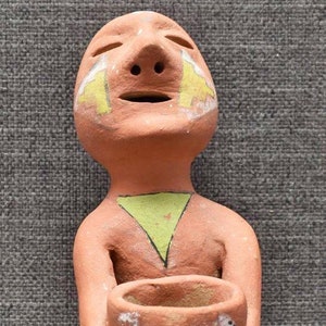 May include: A terracotta figurine of a person with painted details, holding a small terracotta pot with painted designs. The figurine has a closed-eye expression and a painted green triangle on its chest.