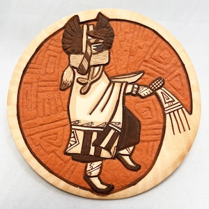 Puede incluir: Un azulejo de cerámica con un diseño pintado a mano de una figura estilizada que lleva una túnica blanca y un tocado. La figura sostiene un objeto largo y delgado en su mano derecha. El azulejo está decorado con un fondo marrón y naranja.