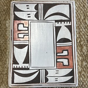 May include: A white ceramic tile with a geometric design in black and brown. The tile has a central rectangular area with a white background. The tile is decorated with a traditional Southwestern design.