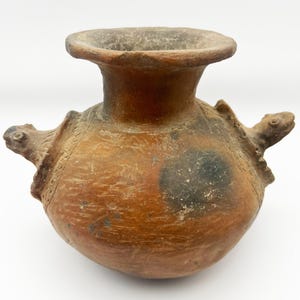 May include: A terracotta pottery vase with two turtle-shaped handles. The vase has a wide mouth and a rounded body. The surface is brown with black markings.