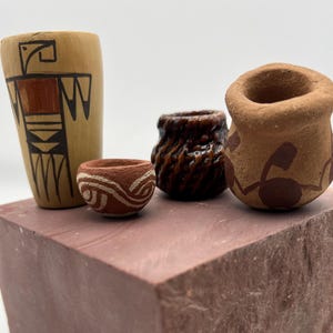 May include: Four small, handcrafted pottery vessels of varying shapes and designs. One is a tall, light-colored vase with a bird figure. Another is a small, round pot with a white swirl pattern. The other two are brown and tan with unique textures and patterns.
