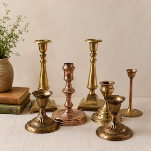May include: A collection of antique brass and copper candlesticks of varying heights and designs. The candlesticks are arranged on a light-colored surface, with a vase of greenery and books in the background. The candlesticks have a vintage aesthetic.