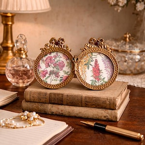 Puede incluir: Un marco de fotos dorado de doble cara con estampados florales. El marco tiene un diseño de lazo en la parte superior y está sobre una pila de libros antiguos. También se ven un bolígrafo dorado y una pulsera de perlas.