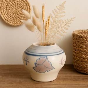 Può includere: Un vaso in ceramica color crema con dettagli blu e fiori secchi. Il vaso è posto su una superficie di legno. Sullo sfondo sono visibili un sottobicchiere e un cestino. Il vaso è alto circa 15 cm.