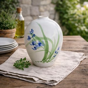 May include: White ceramic vase with a rounded shape, hand-painted with blue floral accents and green leaves. The vase is displayed on a wooden table with a linen cloth, showcasing a rustic, natural aesthetic. A bottle of olive oil and a small plant are in the background.
