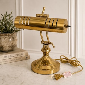 May include: A polished brass banker lamp featuring a cylindrical shade with vertical ventilation slits, an adjustable arm with a central tension knob, and a circular base. The lamp includes a gold power cord with a two-prong plug and a small tag.