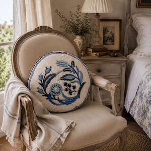 May include: A round, off-white throw pillow featuring intricate blue and navy floral embroidery. The pillow is placed on a light-colored wooden armchair with a beige fabric seat and back, draped with a matching beige throw blanket with blue stripes.