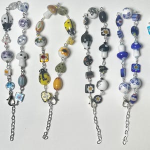 Puede incluir: Una colección de pulseras de cuentas con varios diseños. Las pulseras presentan cuentas coloridas en formas como setas, mariquitas y flores. Cada pulsera tiene una cadena y un cierre plateados.