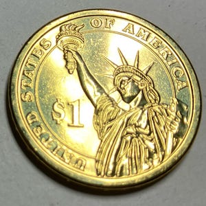 Puede incluir: Una moneda de un d&oacute;lar estadounidense de color dorado. La moneda presenta la Estatua de la Libertad sosteniendo una antorcha, con la inscripci&oacute;n "UNITED STATES OF AMERICA" y "$1". El borde de la moneda es liso.