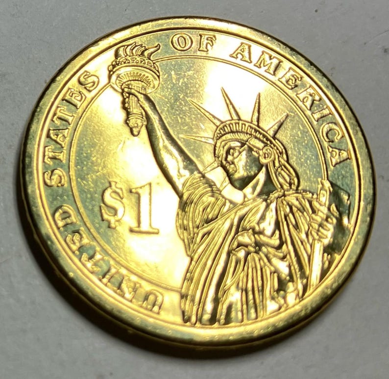 Puede incluir: Una moneda de un d&oacute;lar estadounidense de color dorado. La moneda presenta la Estatua de la Libertad sosteniendo una antorcha, con la inscripci&oacute;n "UNITED STATES OF AMERICA" y "$1". El borde de la moneda es liso.