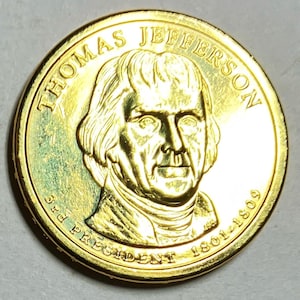 Puede incluir: Una moneda dorada con un retrato detallado de Thomas Jefferson. La moneda est&aacute; inscrita con "THOMAS JEFFERSON", "3rd PRESIDENT" y los a&ntilde;os "1801-1809". La moneda tiene una superficie brillante y reflectante.