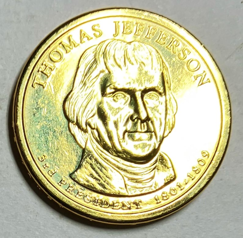 Puede incluir: Una moneda dorada con un retrato detallado de Thomas Jefferson. La moneda est&aacute; inscrita con "THOMAS JEFFERSON", "3rd PRESIDENT" y los a&ntilde;os "1801-1809". La moneda tiene una superficie brillante y reflectante.