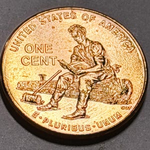 May include: A copper-colored United States one cent coin featuring a relief of a person reading a book while sitting on a log. The text UNITED STATES OF AMERICA, ONE CENT, and E PLURIBUS UNUM are embossed on the circular surface of the coin.