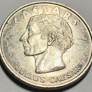 May include: A silver coin featuring a profile of Julius Caesar, encircled by the inscriptions "LAESARS" and "JULIUS CAESAR". The coin's design includes a laurel wreath and detailed facial features.