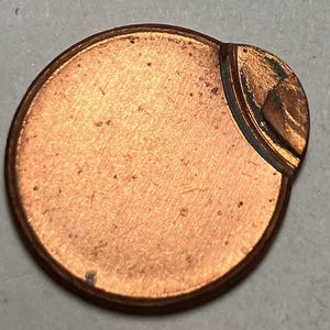 May include: A copper-colored, circular metal object with a unique, curved design on one side. The object has a slightly textured surface and a darker, contrasting edge. The curved section features a raised, detailed pattern.