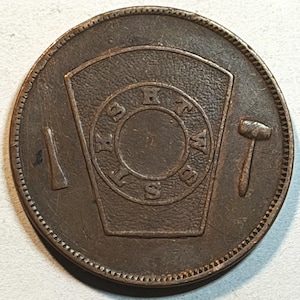 May include: A round, bronze-colored coin with a central design featuring a shield shape and the letters "S B H W S". A hammer and chisel flank the central design. The coin has a textured edge.