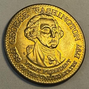 May include: A gold-colored coin featuring a portrait of George Washington. The coin is inscribed with "GEORGE WASHINGTON" and "FIRST PRESIDENT OF THE UNITED STATES", along with the years 1789 and 1797.