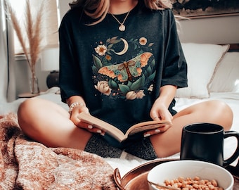 May include: A dark grey t-shirt with a floral design featuring a large butterfly and a crescent moon. The butterfly has orange, green, and brown wings.