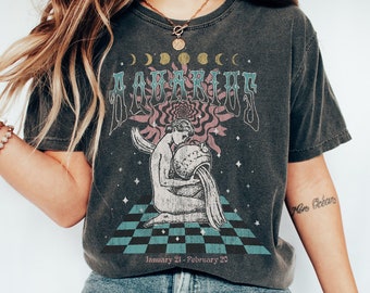 Puede incluir: Una camiseta gris oscuro con un gráfico de estilo vintage del signo astrológico Acuario. El gráfico presenta a una persona vertiendo agua de una jarra, rodeada de estrellas y un patrón de tablero de ajedrez. El texto "Aquarius" y "January 21 - February 20" está impreso en la camiseta.