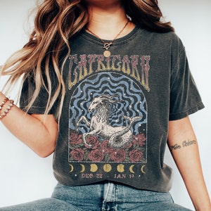 May include: A dark gray t-shirt with a vintage-style graphic of the Capricorn zodiac sign. The graphic features a goat with a fish tail, surrounded by roses and crescent moons. The text "Capricorn" is written in a stylized font above the graphic, and the dates "Dec 22 - Jan 19" are written below it.