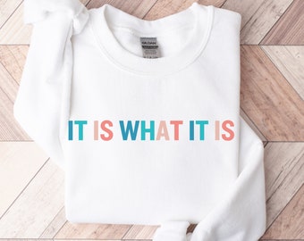 May include: White sweatshirt with the text "IT IS WHAT IT IS" in colorful letters. The letters are in shades of blue, pink, and teal.