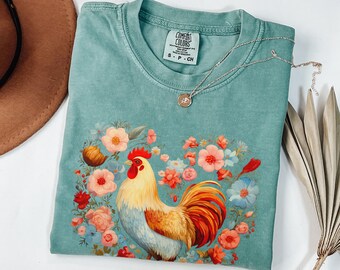 May include: A teal blue t-shirt with a colorful rooster illustration surrounded by flowers. The shirt has a tag that says "Comfort Colors" and "S-P-CH".