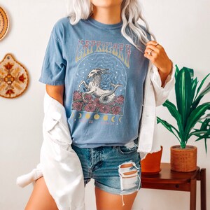 May include: A blue t-shirt with a graphic of a sea goat with stars and moons. The text "Capricorn" is printed on the shirt. The model is wearing the shirt with a white jacket and denim shorts.