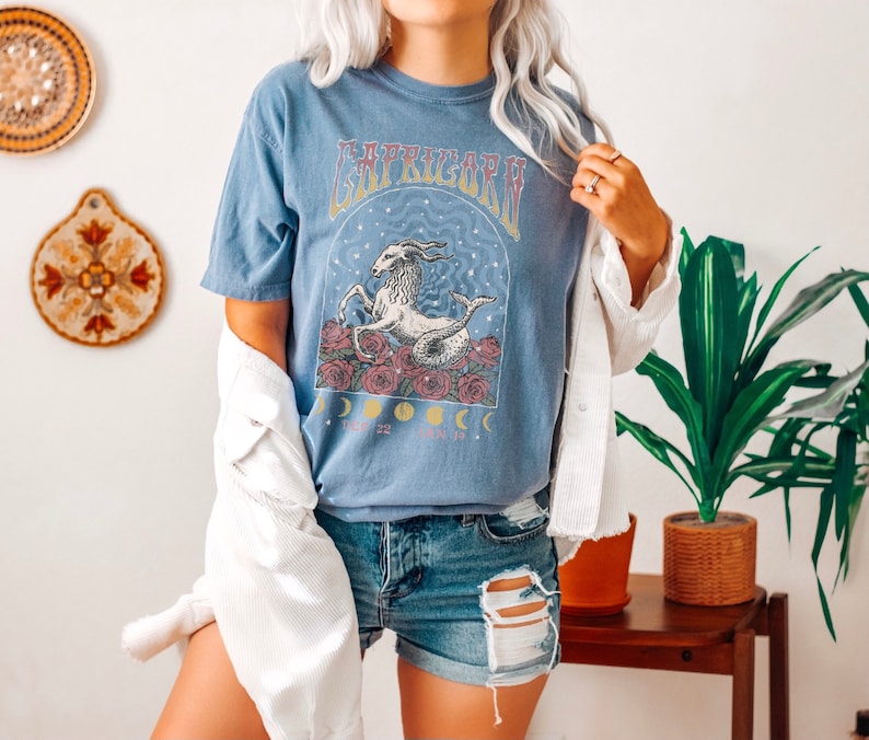 May include: A blue t-shirt with a graphic of a sea goat with stars and moons. The text "Capricorn" is printed on the shirt. The model is wearing the shirt with a white jacket and denim shorts.