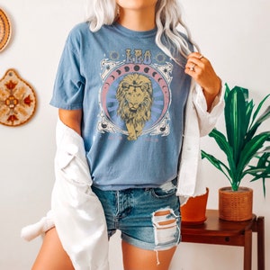 May include: A blue t-shirt featuring a graphic of a lion and the word "Leo" in a retro style. The shirt is being worn by a person with a white denim jacket and denim shorts.